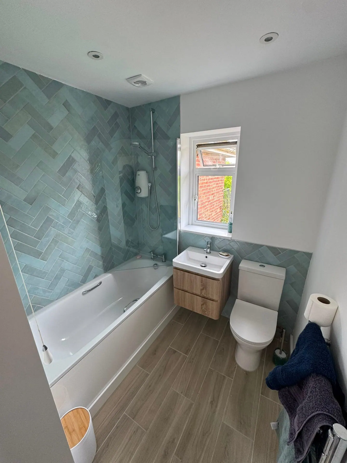 Compact family bathroom with pale blue herringbone tiles, bath shower and floating vanity by Heat Solution London