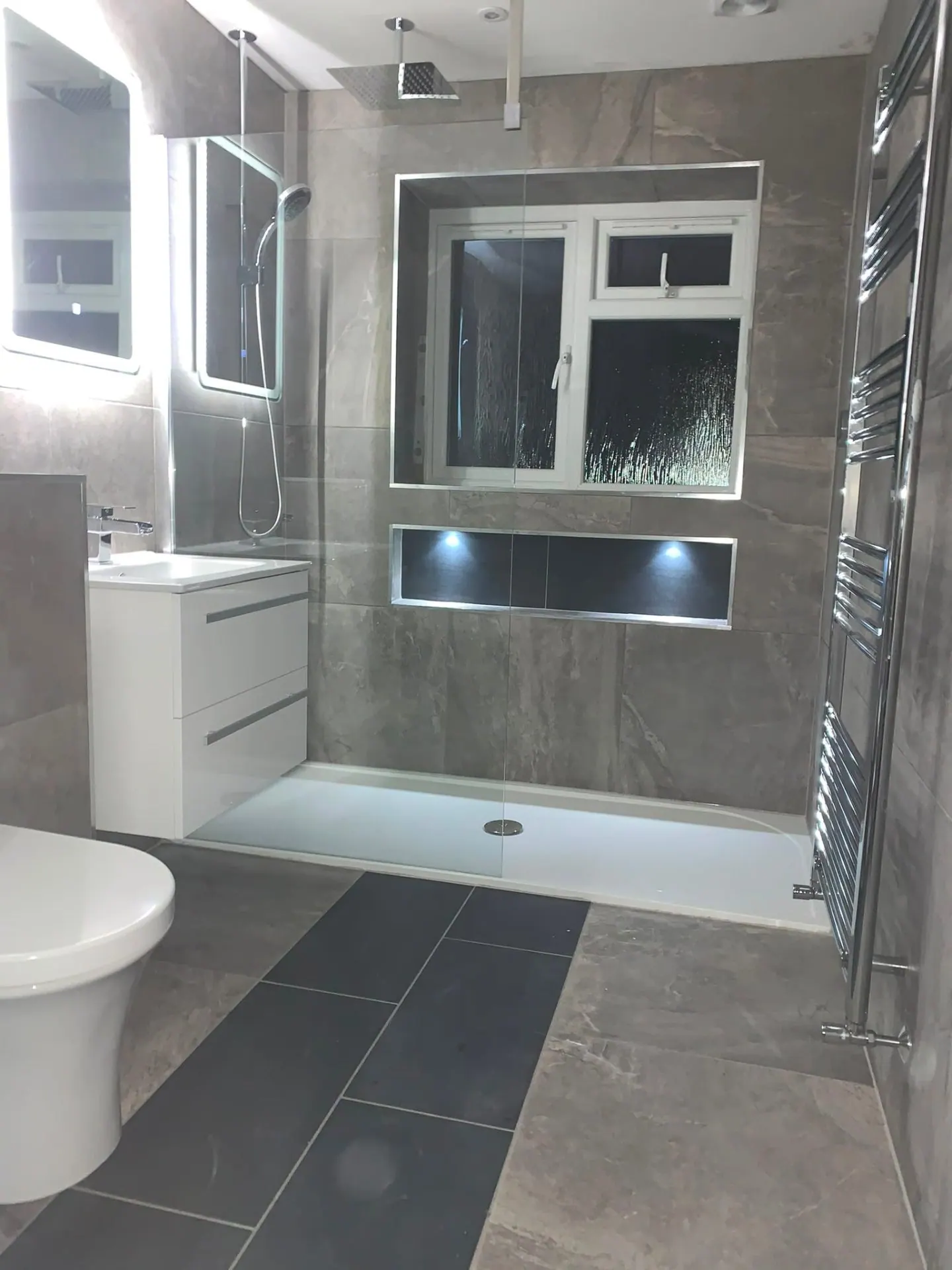 Grey stone wet room with dual rainfall showers, illuminated niche and chrome towel rail by Heat Solution London