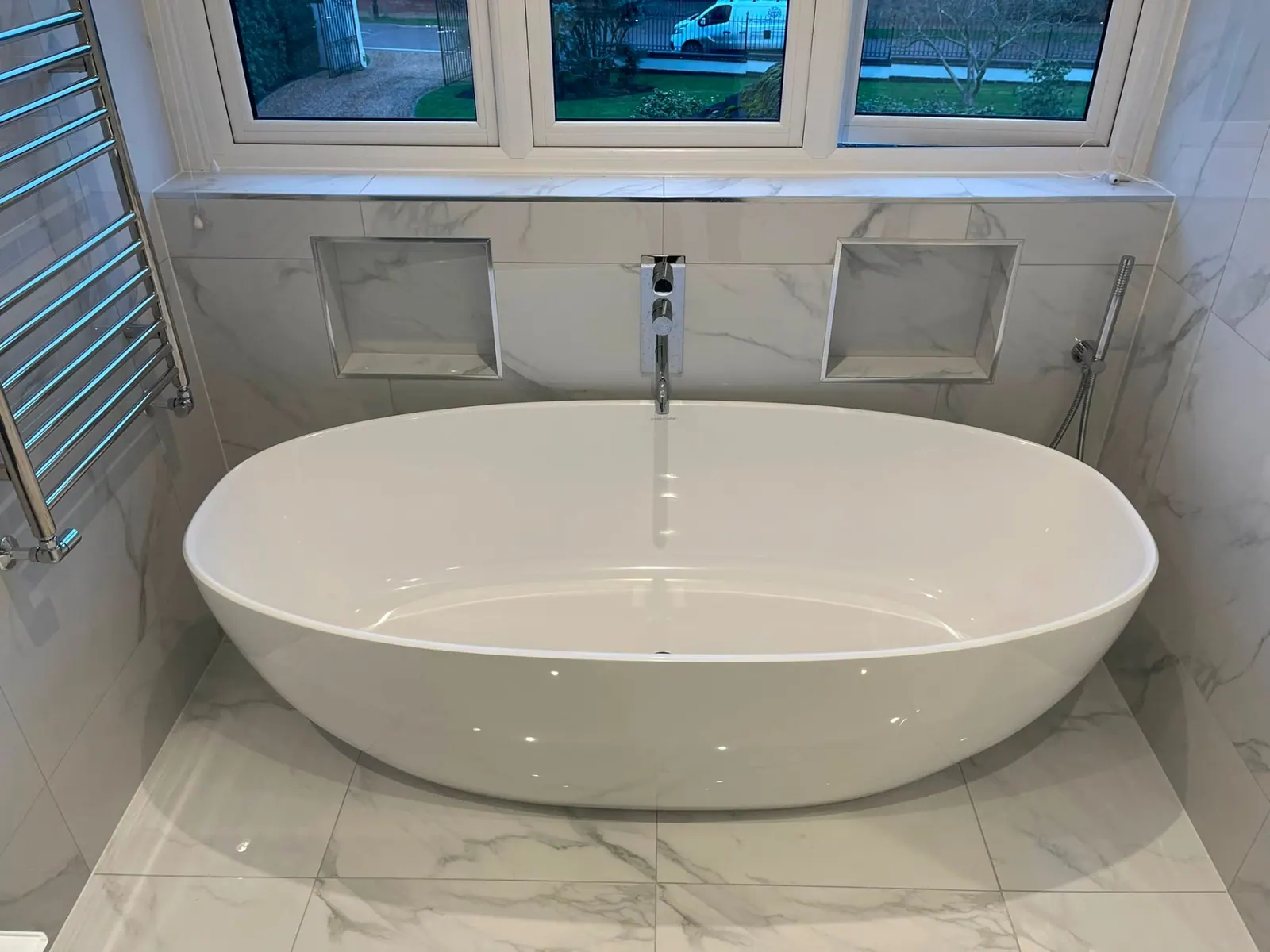 Freestanding oval bath beneath a window in a marble bathroom by Heat Solution London