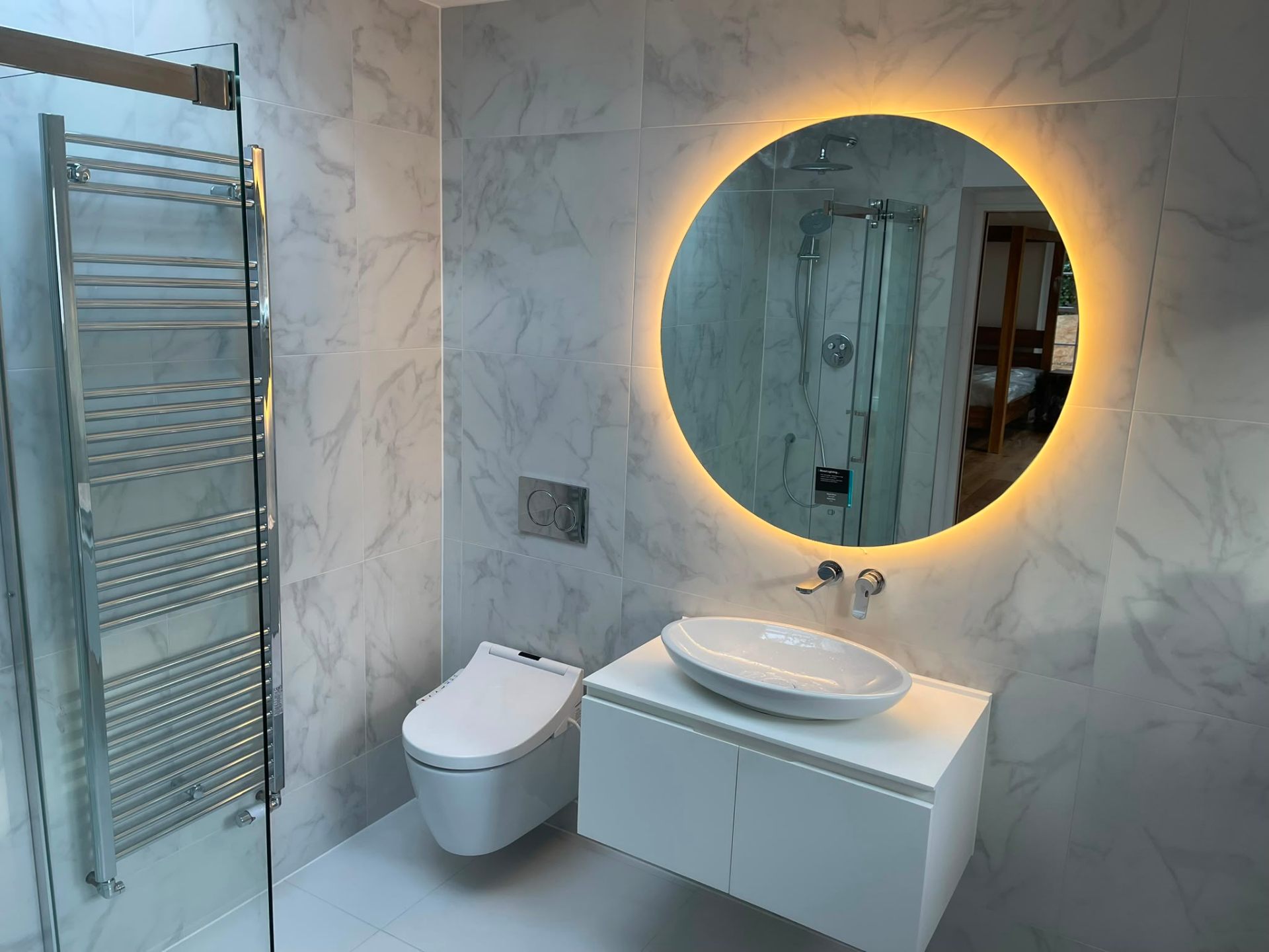 Contemporary bathroom installation with backlit round mirror, wall-hung WC and towel rail in Surrey